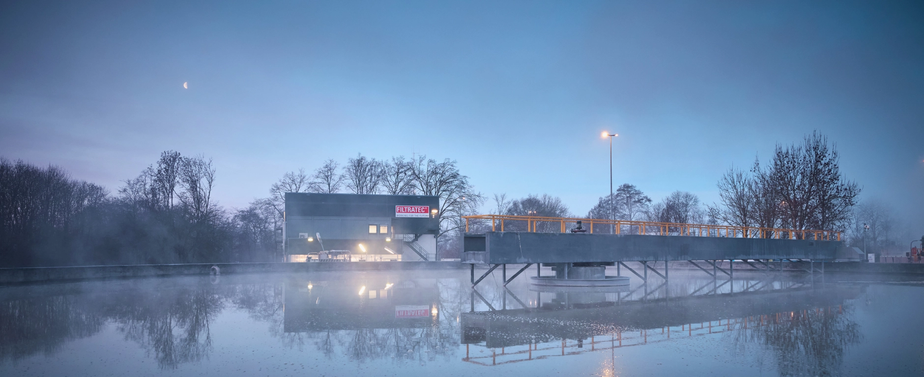 Nebelige Industrieanlage an einem Wasserbecken bei Morgendämmerung, mit beleuchtetem Gebäude mit FILTRATEC-Logo, Stegkonstruktion und Spiegelung im ruhigen Wasser, Mond am Himmel.