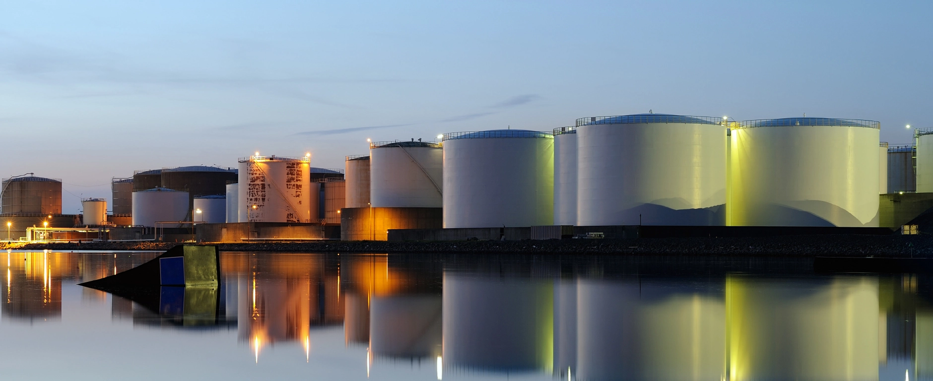 Große zylindrische Industrietanks an einem Hafen bei Dämmerung, beleuchtet und gespiegelt in der ruhigen Wasseroberfläche.