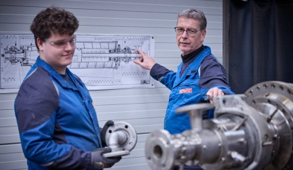 Ein Meister erklärt einem Auszubildenden in einer Werkstatt ein metallisches Anlagenteil anhand einer technischen Zeichnung.